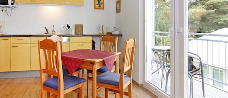 A dining area with a wooden table and chairs, a yellow kitchen in the background, and a balcony with outdoor furniture.