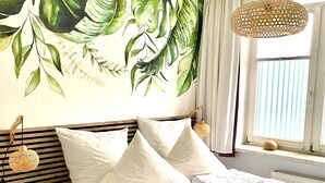 A bedroom with a large bed, white bedding, a wooden headboard, a small bedside table, a woven lamp, and a large botanical mural on the wall. - Ferienwohnung Strandlaufer in Zinnowitz (Zinnowitz)