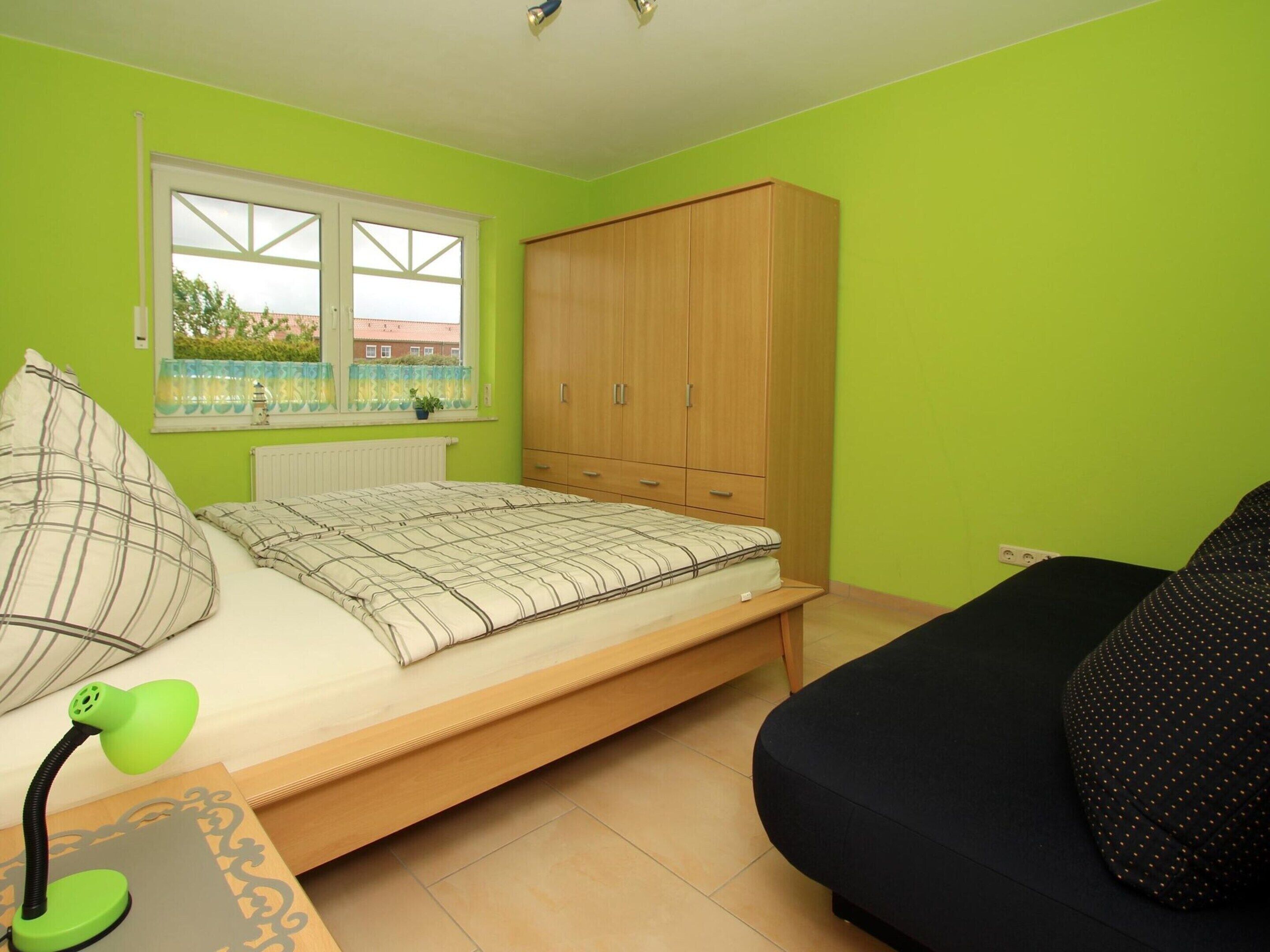 A bedroom with a bed, a wardrobe, a desk with a lamp, and a window with a view of a building and trees.