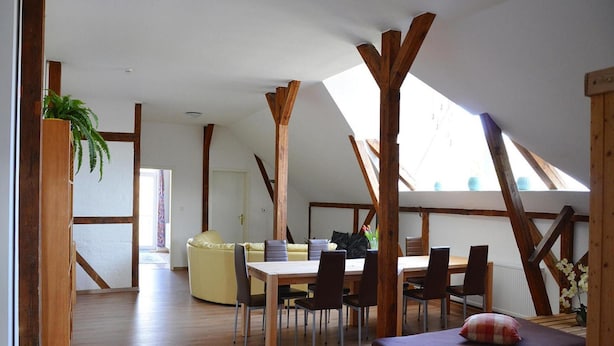A spacious loft bedroom with a high ceiling, wooden beams, a skylight, a bed, a dining table with chairs, and a sofa. - Ferienwohnung an der Elde (Luebz)
