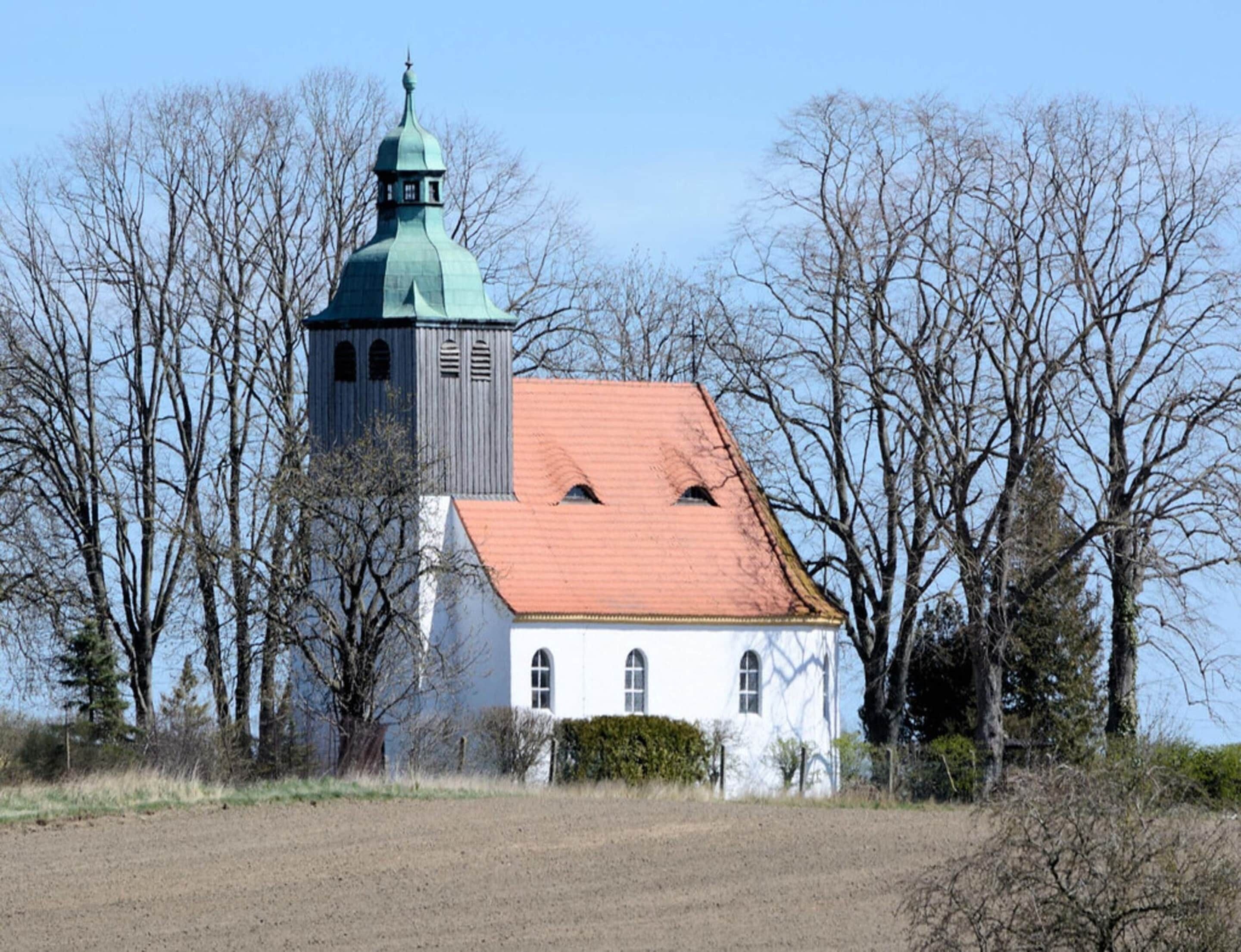 A white building with a red roof and a green tower, surrounded by leafless trees.