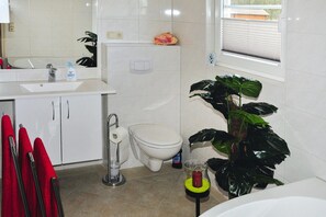 A bathroom with a white sink, toilet, and bathtub. There are red towels, a plant, and a glass jar with a brush.