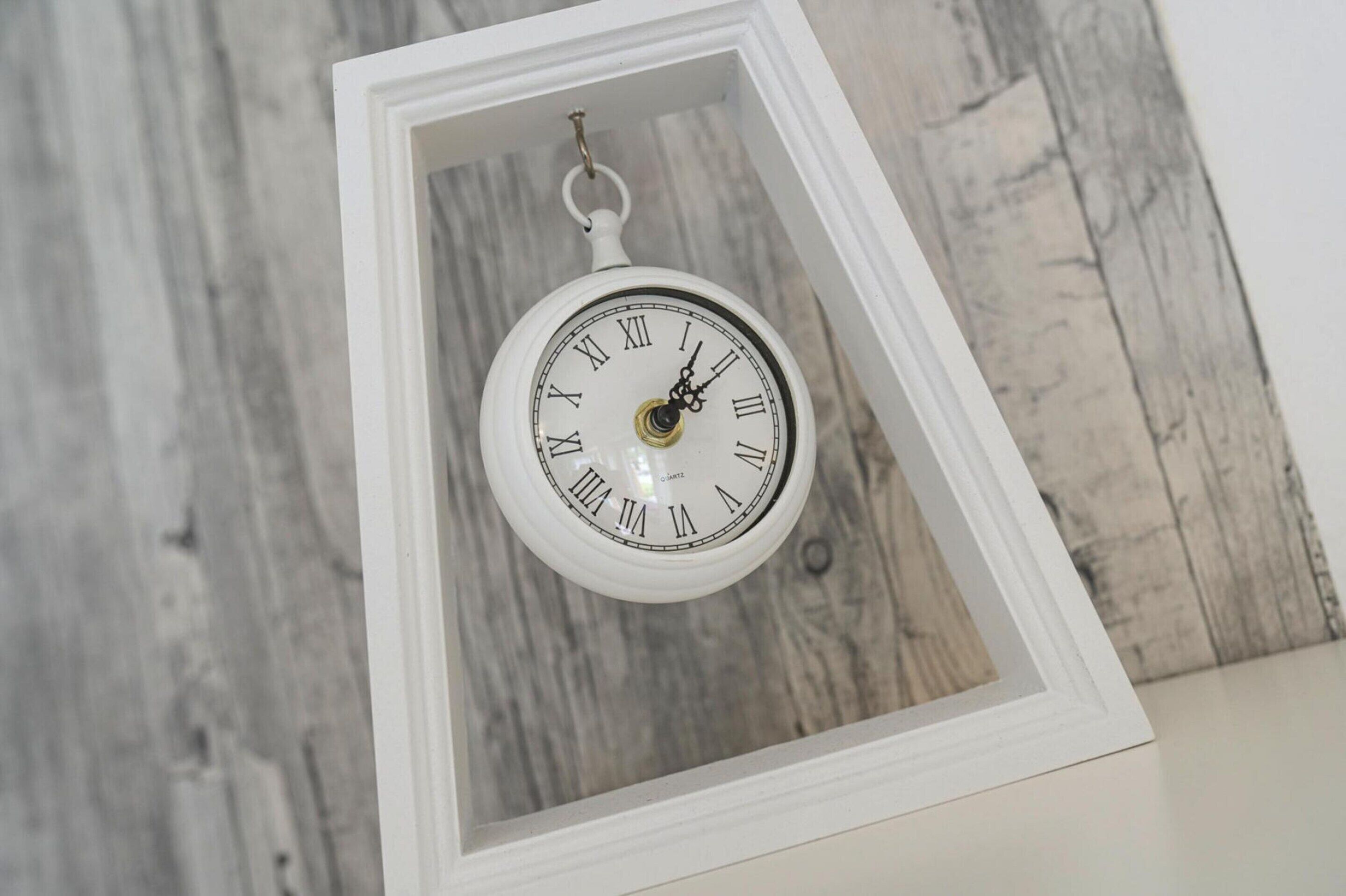 A white clock with Roman numerals in a white frame against a wooden background.