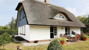 A thatched-roof house with a large window, a patio with chairs, and potted plants. - Ferienwohnung, Wieck am Darss (Wieck auf dem Darss)