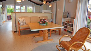 A cozy living room with a wooden dining table, a sofa with patterned cushions, a wicker rocking chair, and a bookshelf. - Ferienhaus, Krusendorf (Schwedeneck)
