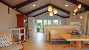 A spacious room with a wooden floor, a dining table with a flower centerpiece, a sofa with a polka dot pillow, and a bookshelf. - Ferienhaus, Krusendorf (Schwedeneck)