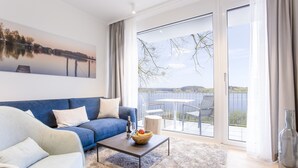A modern living room with a blue sofa, a coffee table with fruit, and a large window overlooking a balcony with outdoor furniture.