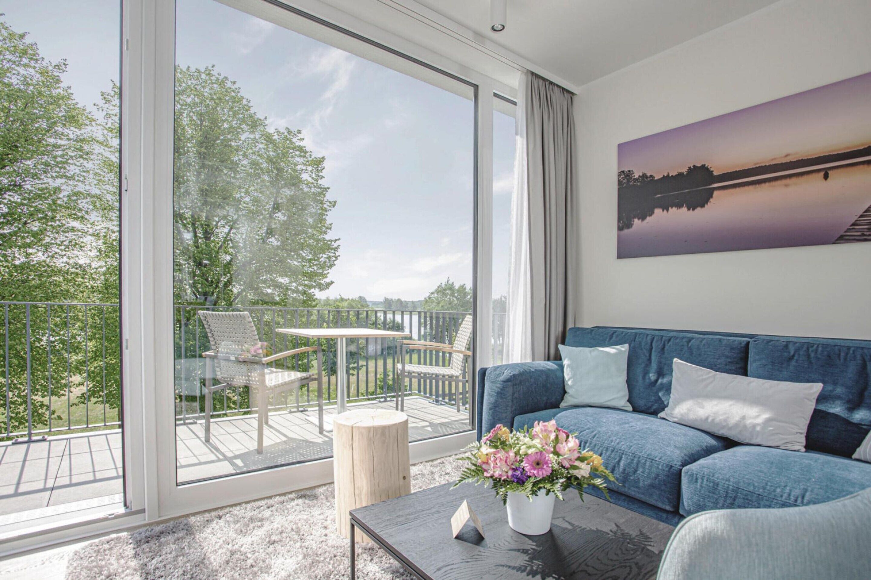 A modern living room with a large sliding glass door leading to an outdoor balcony. The room features a blue sofa, a coffee table with a vase of flowers, and a painting of a sunset over a lake.