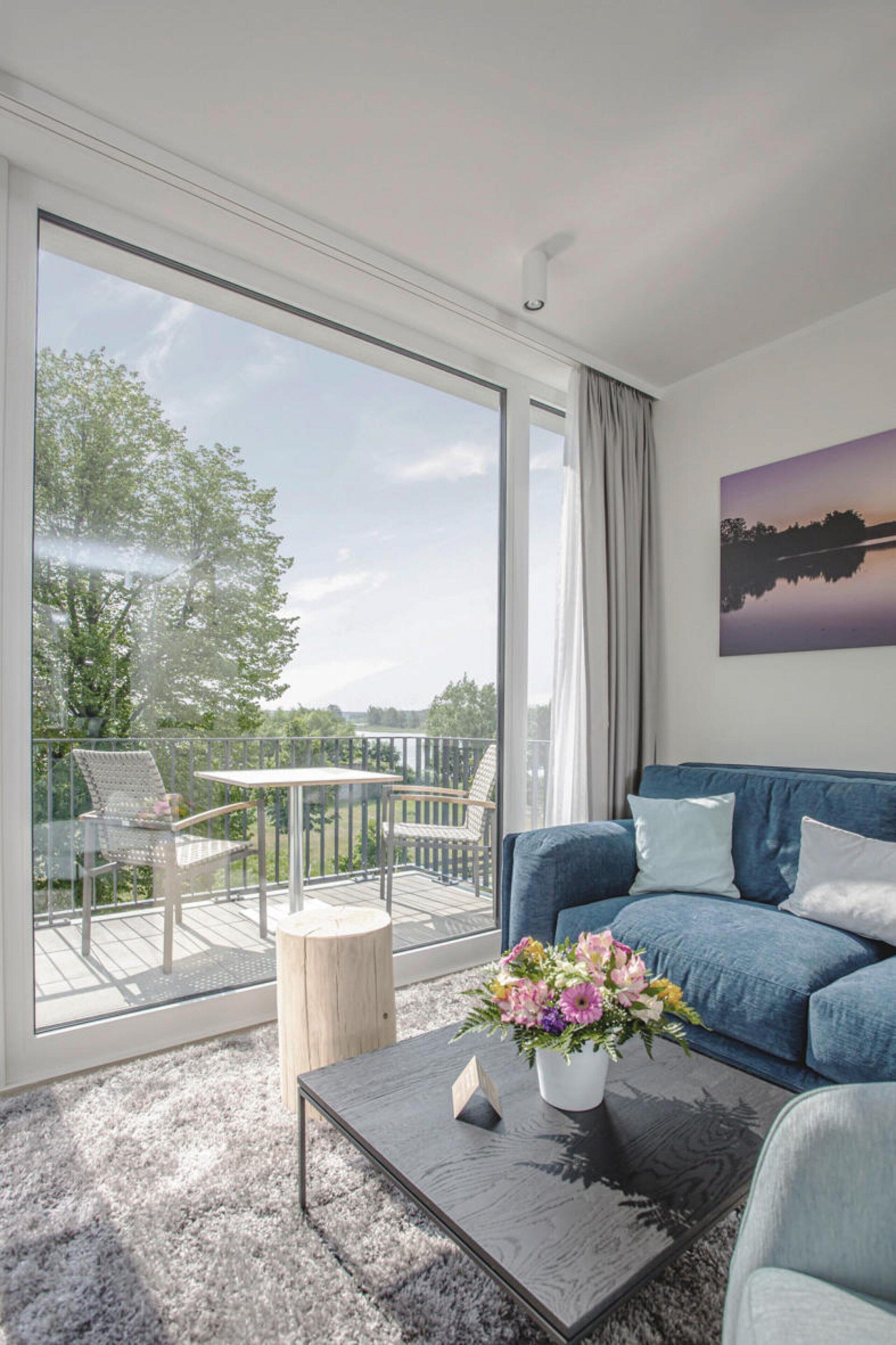 A modern living room with a large sliding glass door leading to an outdoor deck, a blue sofa, a coffee table with a vase of flowers, and a painting on the wall.