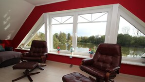 Ein Zimmer mit schrägem Dach, einem großen Fenster mit Seeblick und zwei Ledersesseln mit passenden Hocker.
