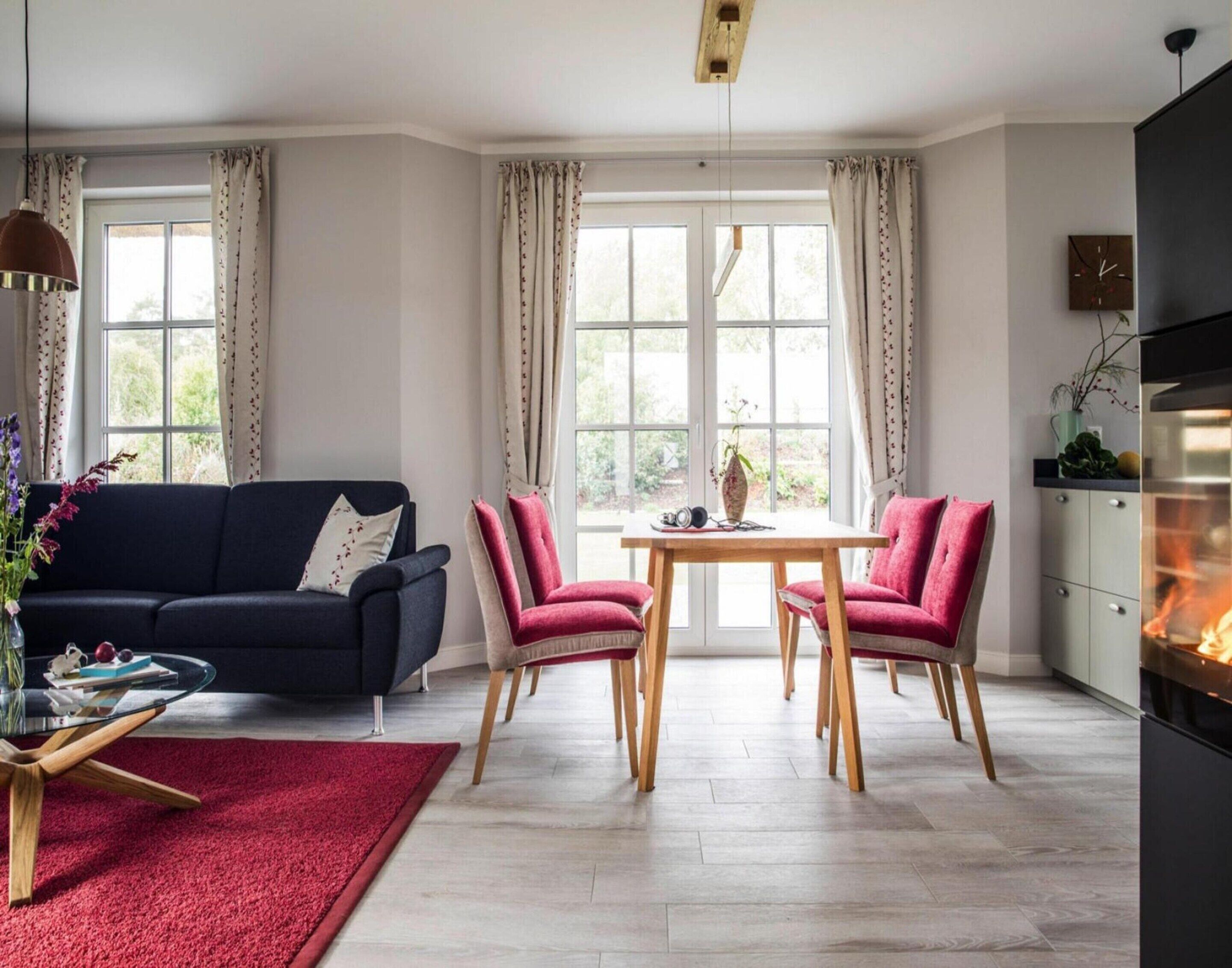 Ein modernes Wohnzimmer mit einem dunkelblauen Sofa, zwei roten Sesseln, einem Holztisch und einem roten Teppich. Rechts befindet sich ein Kamin und ein großes Fenster mit Vorhängen.