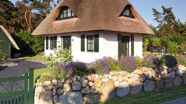 Una casa con techo de paja, paredes blancas y contraventanas verdes, rodeada por un muro de piedra y plantas en flor.