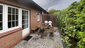 Une maison en briques dotée d’un patio comprenant une table et des chaises en bois, une antenne parabolique et un jardin à la végétation luxuriante.