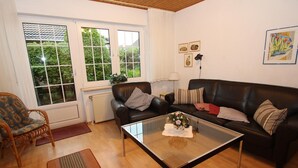 Un salon avec une table basse en verre, deux canapés en cuir, un fauteuil en osier et une vue sur la verdure grâce à de grandes fenêtres.