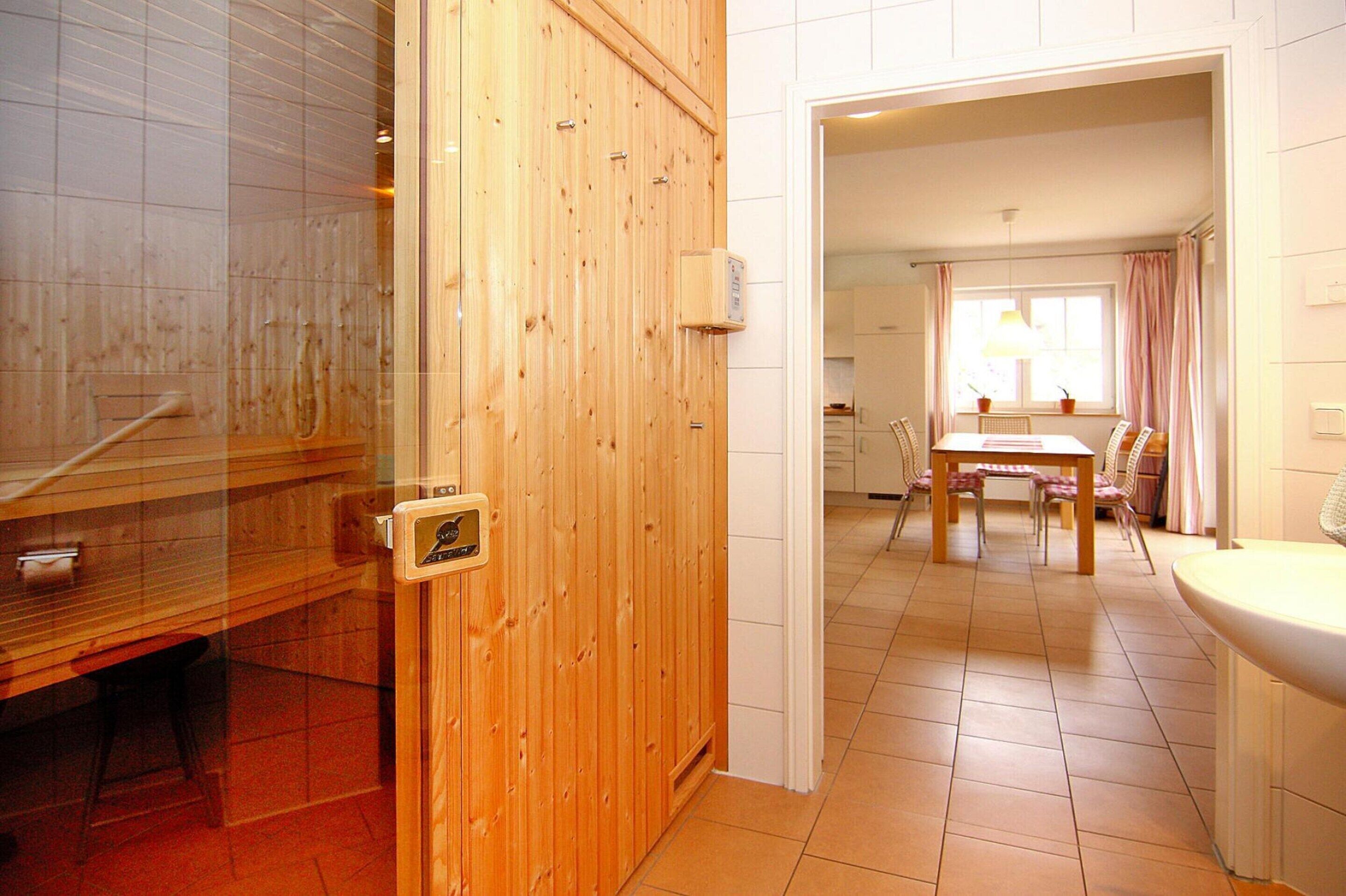 Una porta in legno che dà su una stanza con un tavolo e sedie, una finestra con tende, e un lavandino bianco.