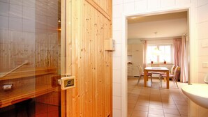 Une porte en bois donne sur une pièce où se trouvent une table à manger et des chaises, une fenêtre avec des rideaux et un évier blanc.