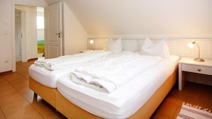Un lit bien fait dans une pièce dont le plafond est en pente, une table de chevet avec une lampe, et une vue sur un paysage coloré par une porte ouverte.