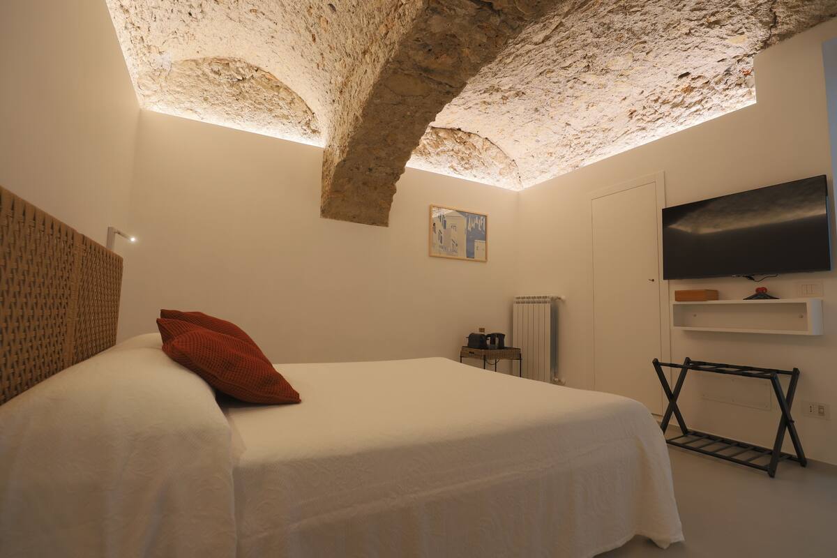 A bedroom with a bed, a TV, a radiator, and a stone ceiling.