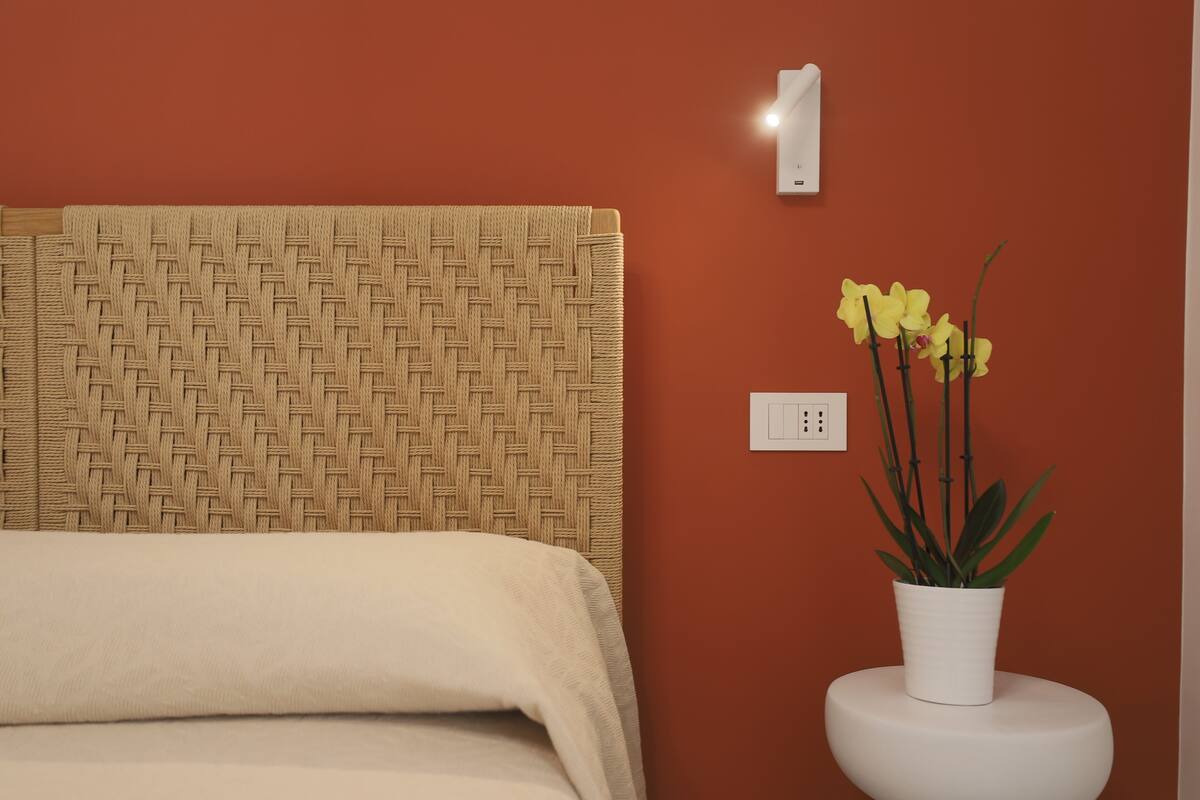 A bed with a woven headboard, a white bedside table with a potted plant, and a wall socket.