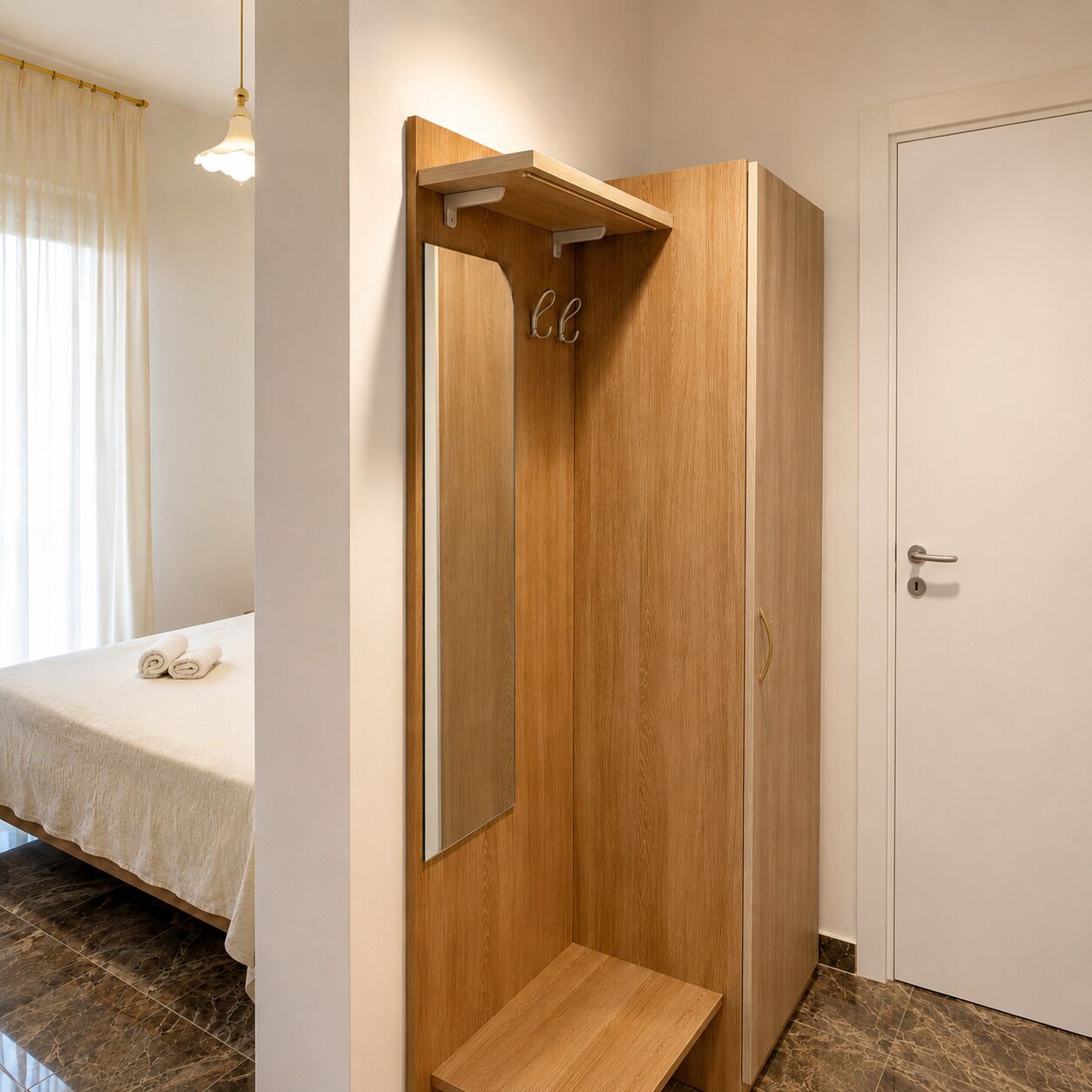 A hotel room with a wooden wardrobe, a small wooden bench, a bed with a white bedspread, and a white door.