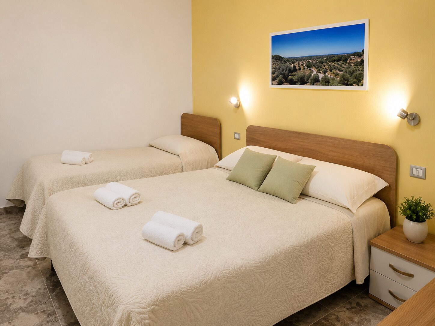 Habitación de hotel con dos camas, una mesita de noche con lámpara, un cuadro en la pared y una pequeña planta en maceta.