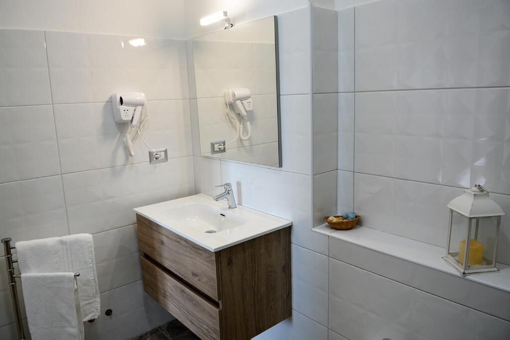 A modern bathroom with a white sink, wooden vanity, and a mirror. There are two hair dryers mounted on the wall, a towel rack, and a small shelf with a candle and a lantern.