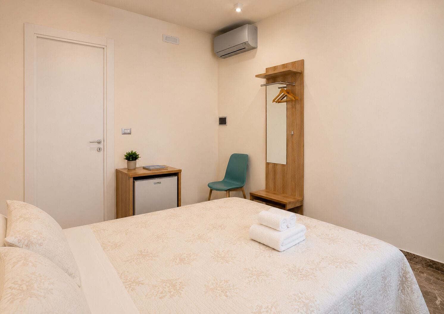 A hotel room with a bed, a desk, a chair, a mirror, and a small plant.