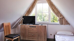 A bedroom with a sloped ceiling, a wooden desk with a TV, a chair, and a bed with white bedding. - Ferienhaus Nachtigall, Glowe (Glowe)