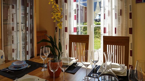 A dining area with a table set for four, including wine glasses and plates, a vase with yellow flowers, and a view of the outside through large windows with patterned curtains.