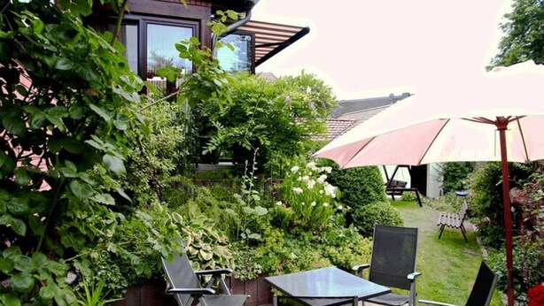 Une terrasse extérieure avec une table et des chaises, entourée d’une végétation luxuriante et protégée par un grand parasol.