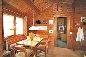 A wooden cabin interior with a dining table, chairs, a bench, a hanging light fixture, and a doorway leading to another room.