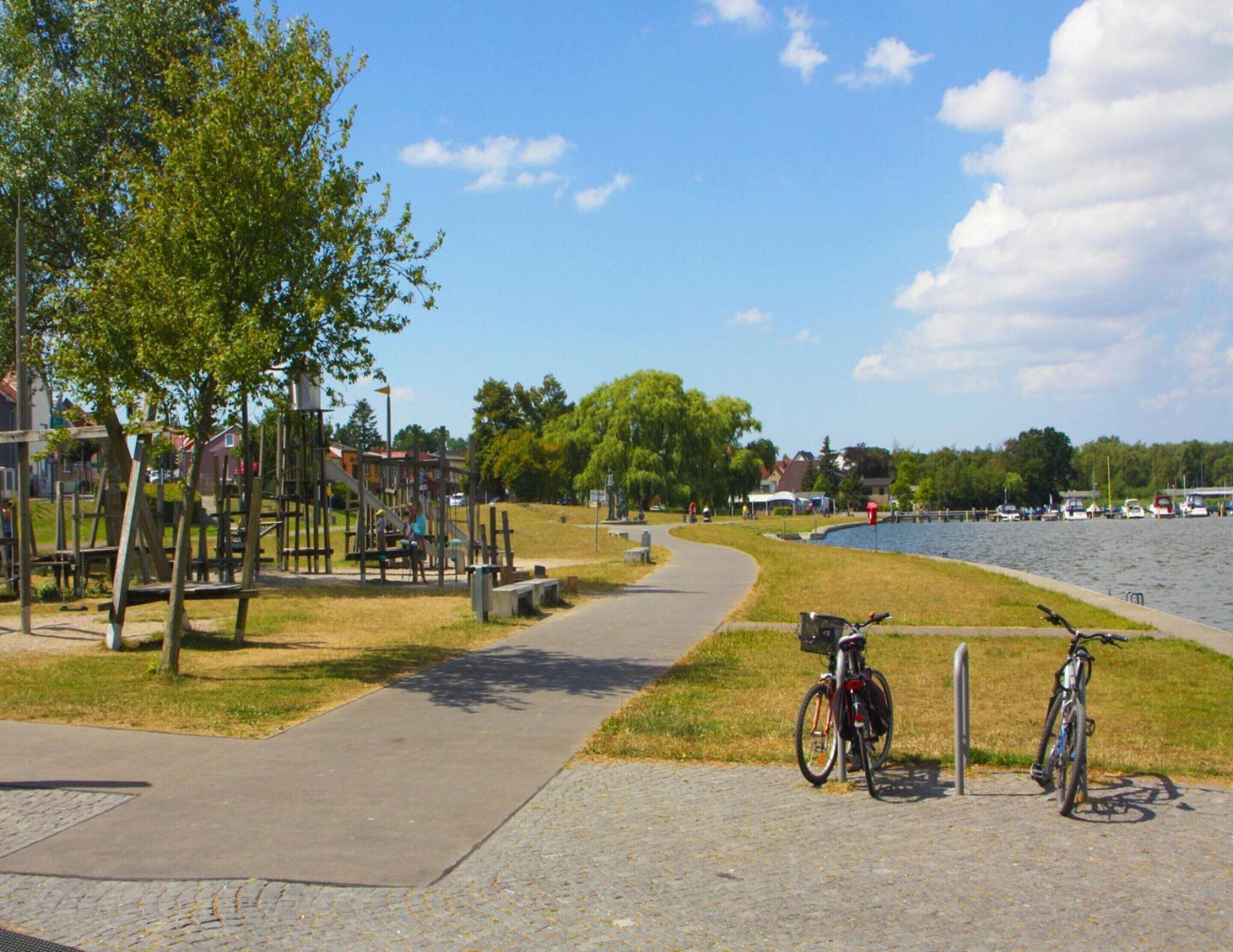 A park with a paved path, playground, and bicycles parked by the water.