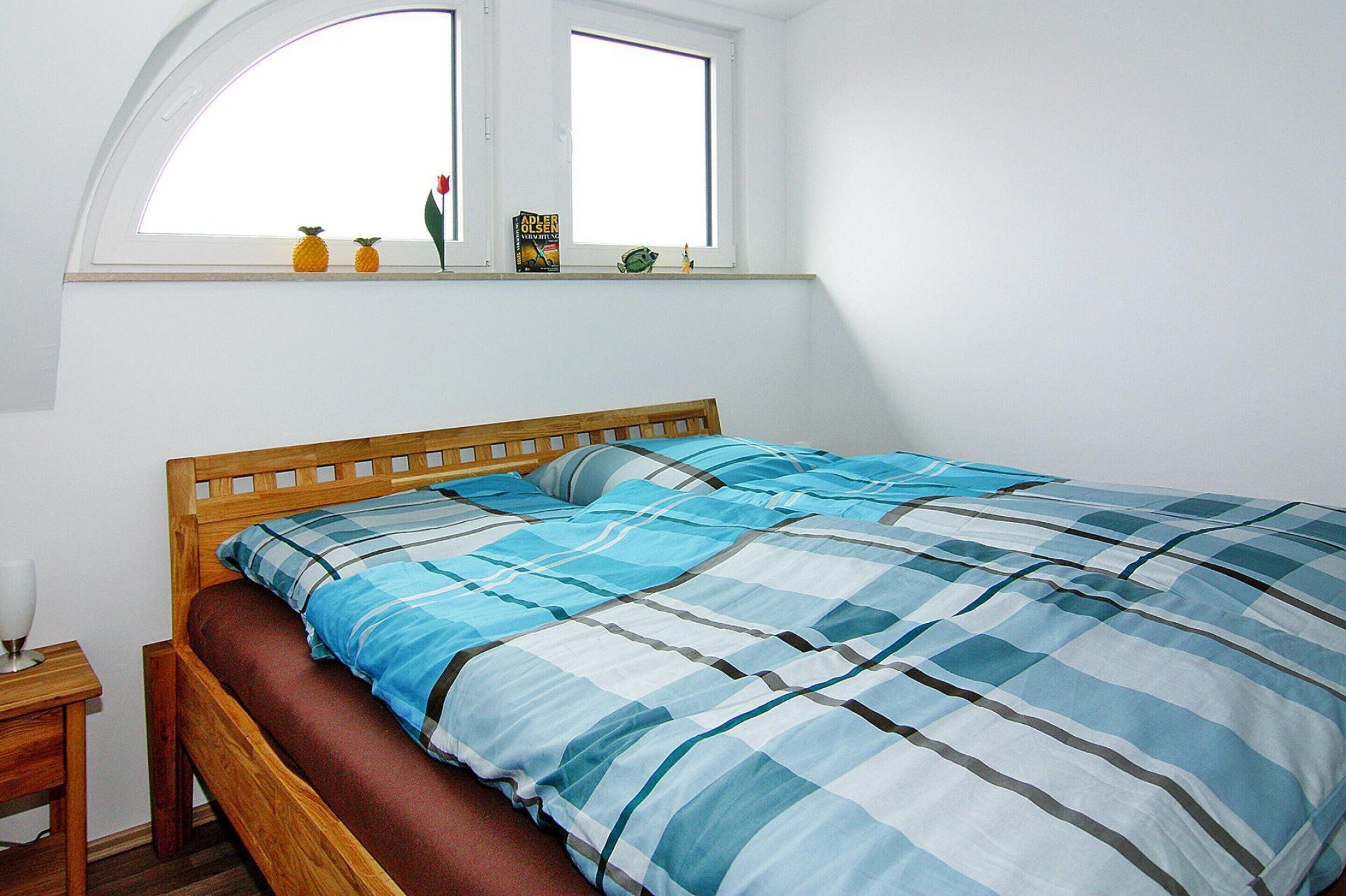 Ein Schlafzimmer mit einem hölzernen Bett, einer blau-weißen Streifen-Bettdecke, einem kleinen Fenster mit zwei Büchern und einer Blume im Fensterbrett sowie einem Nachttisch mit einer Lampe.