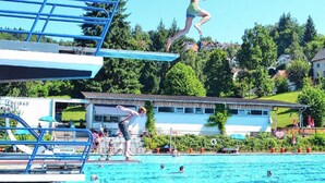Une piscine dotée d’une plateforme de plongeon, de sièges au bord de la piscine et d’un bâtiment en arrière-plan.