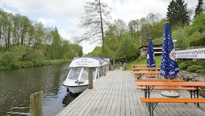 Ein Holzsteg, am Ende liegt ein Boot mit dem Namen "Café Am Brunnen Pension". Auf dem Steg befinden sich Picknicktische und Sonnenschirme.