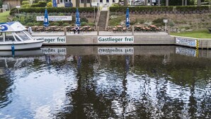 Ein Uferbereich mit einem angedockten Boot, Außenbereich mit Sonnenschirmen und einem Schild mit der Aufschrift 'Wasserwanderplatz'.