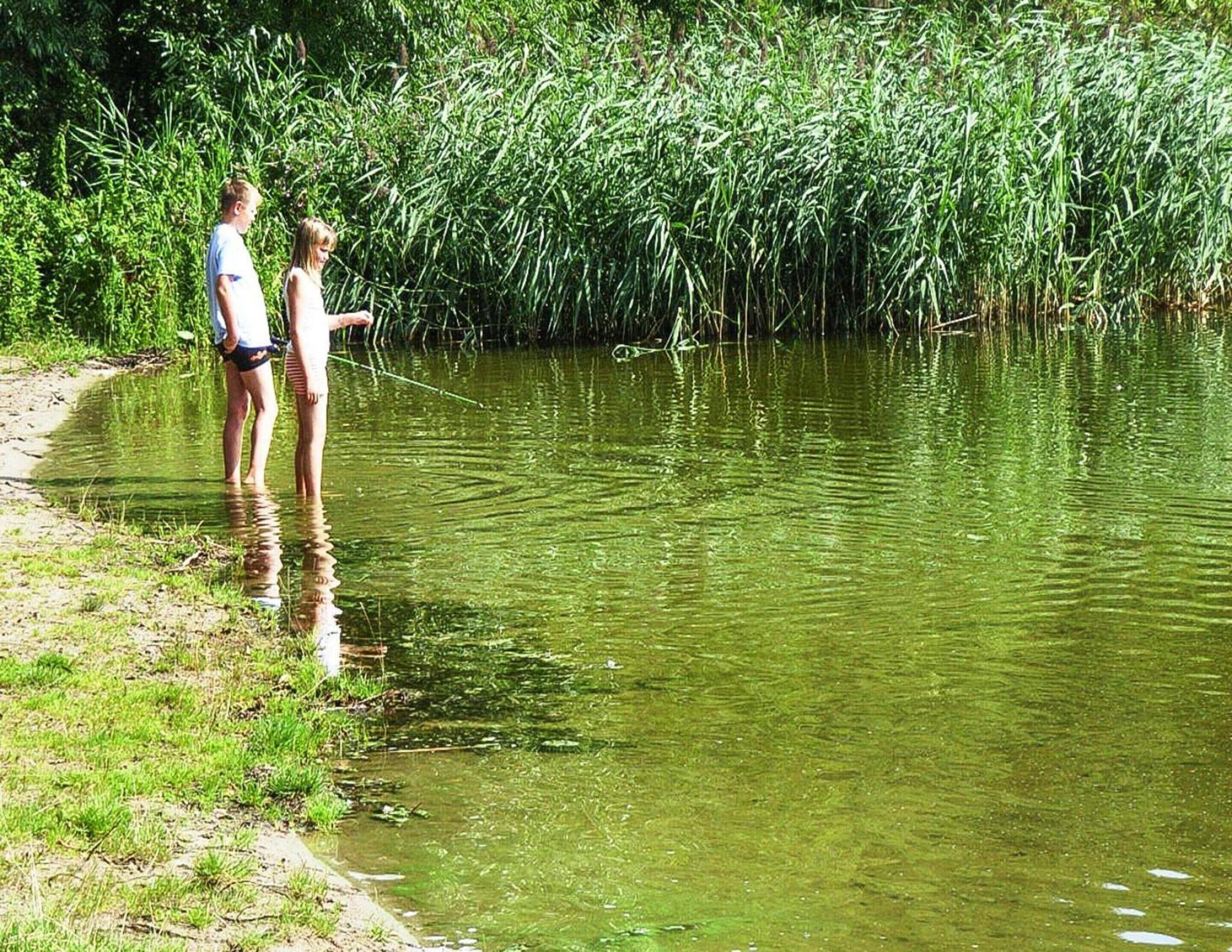 Two people standing by a calm river with dense vegetation in the background.