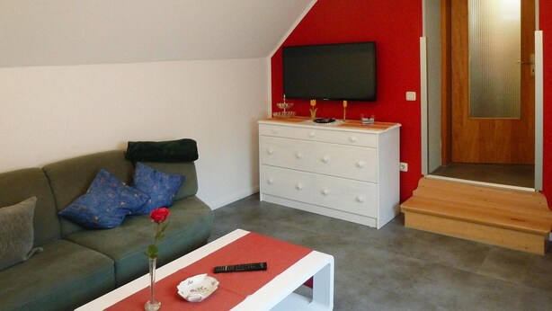 Een woonkamer met een groene bank, een witte salontafel met een rode kleed, een witte dressoir, en een televisie aan de muur.