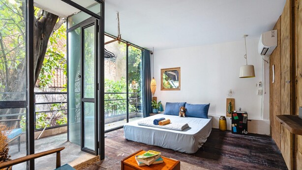 A bedroom with a large bed, a wooden table with books, a window with a view of greenery, and a wooden wall. - HoLo Bato SaiGon - Serviced HomeStay (Ho Chi Minh City)