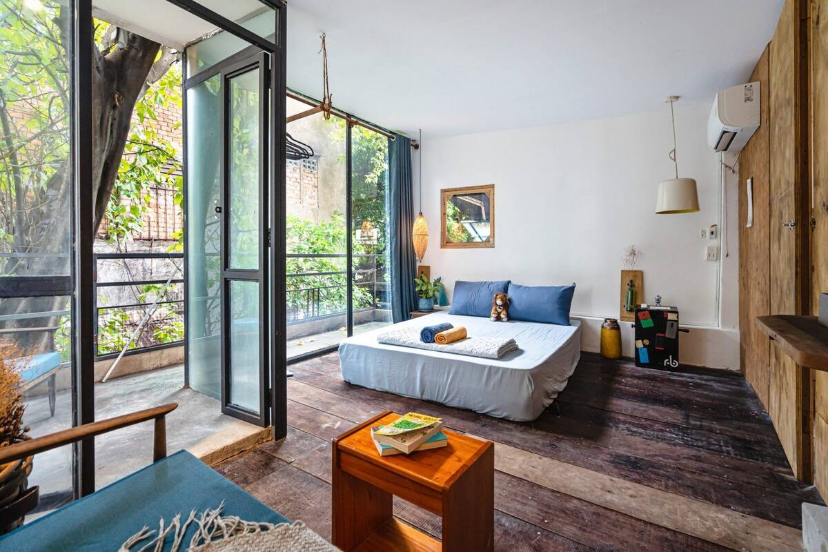 Une chambre avec un grand lit, une table en bois sur laquelle sont posés des livres, une fenêtre donnant sur un paysage verdoyant et un mur en bois.