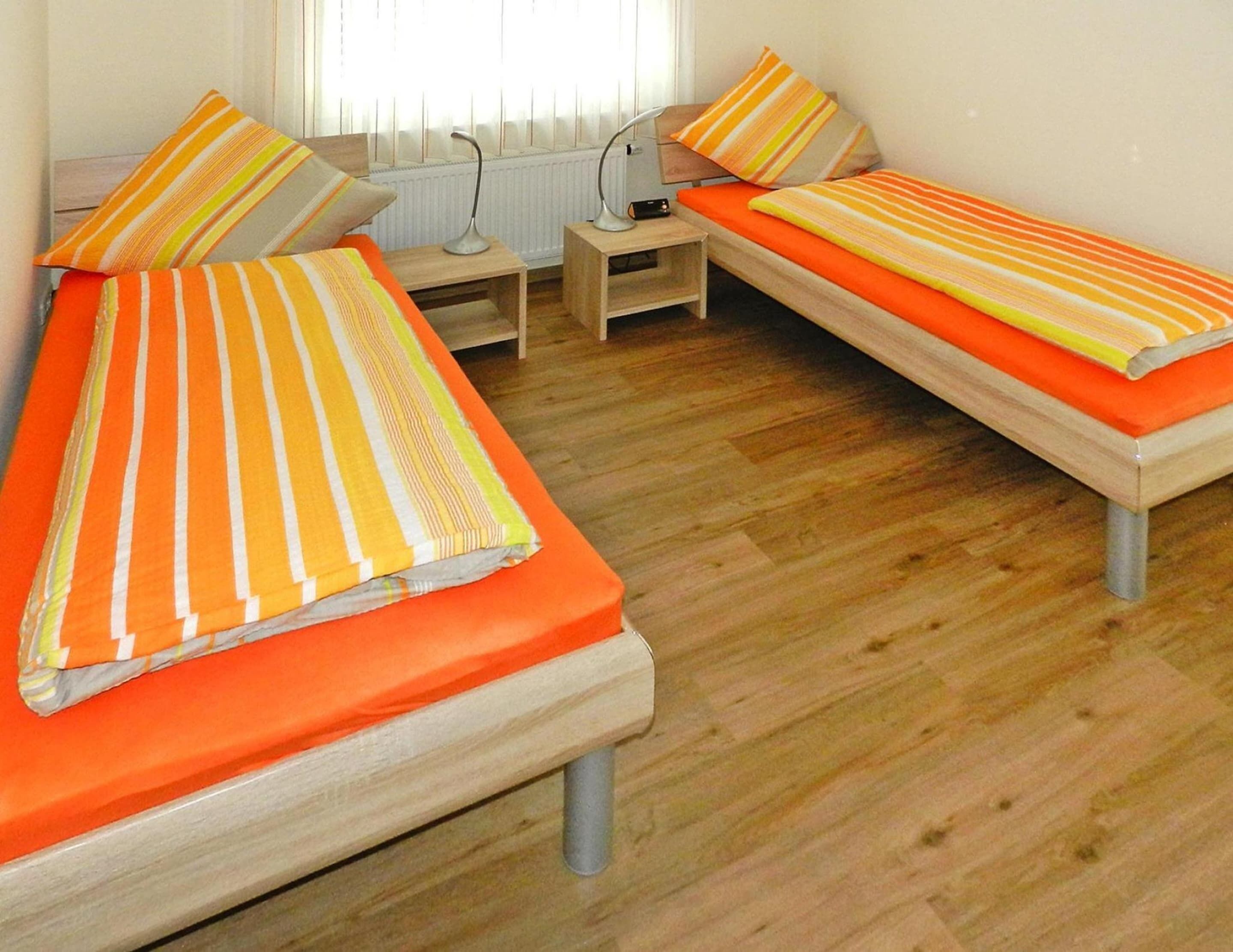 A room with two single beds, each with striped bedding and orange pillows. There is a small wooden nightstand with a lamp between the beds.