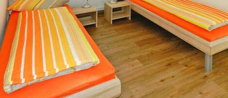 A room with two single beds, each with striped bedding and orange pillows. There is a small wooden nightstand with a lamp between the beds.