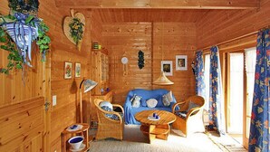 Ein gemütliches Holzhaus-Interieur mit einem blauen Sofa, Korbstühlen, einem kleinen runden Tisch und einem großen Fenster mit floralen Vorhängen.