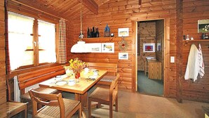 Ein Holzblockhaus-Innenraum mit einem Esstisch, gedeckt für vier Personen, einer Bank und einem durch einen offenen Durchgang sichtbaren Küchenbereich.