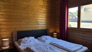 En stuga med träpanel, ett rum med en säng, två sänglampor och ett fönster med utsikt över hus.