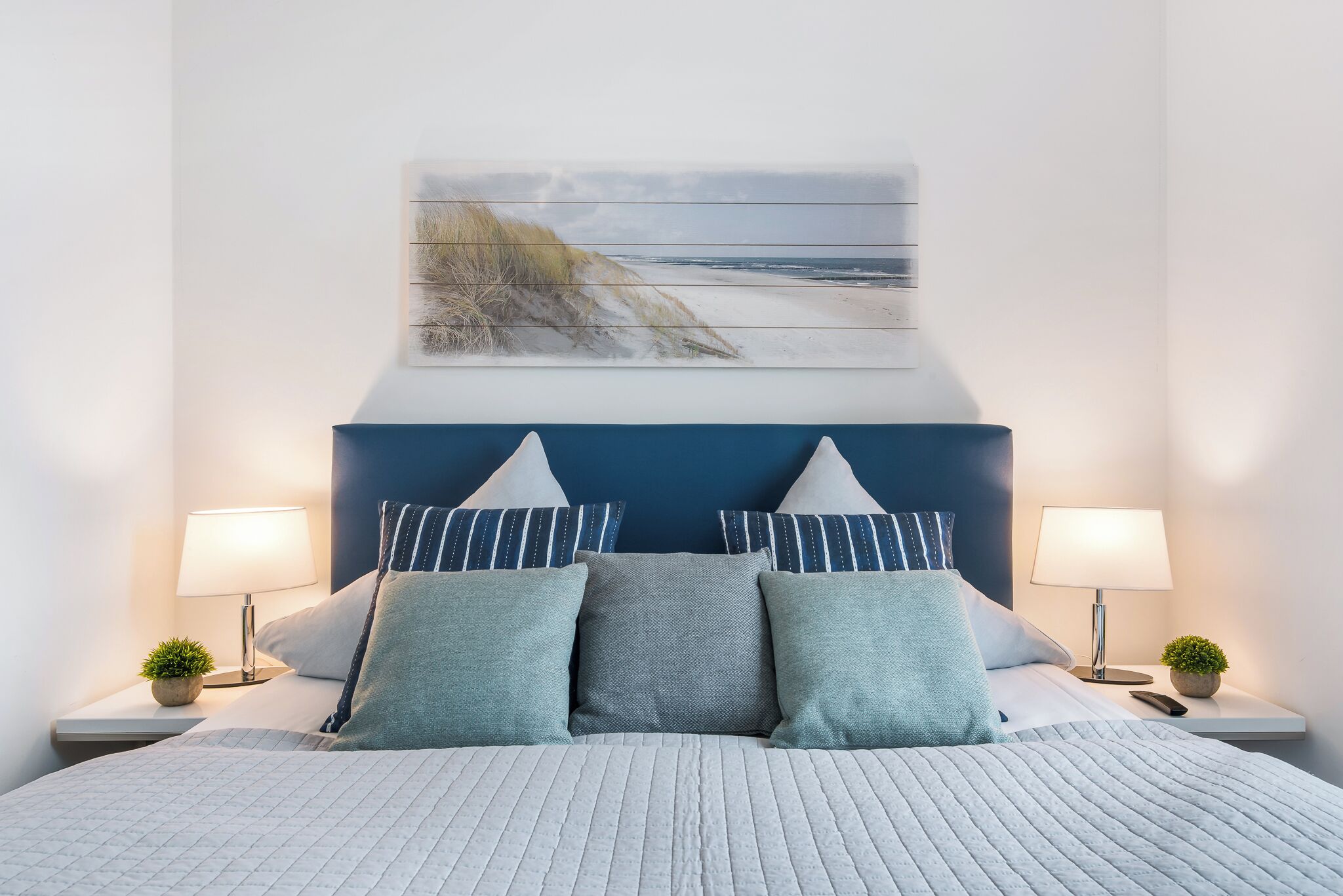 Un lit bien fait, agrémenté d’un coussin à rayures, d’une tête de lit ornée d’une œuvre d’art évoquant le bord de mer, et de lampes de chevet avec de petites plantes en pot.