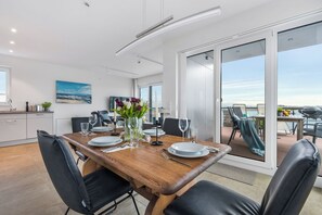 Un espace repas moderne avec une table en bois dressée pour quatre personnes, des chaises noires et une vue sur l’extérieur grâce à de grandes portes-fenêtres coulissantes.