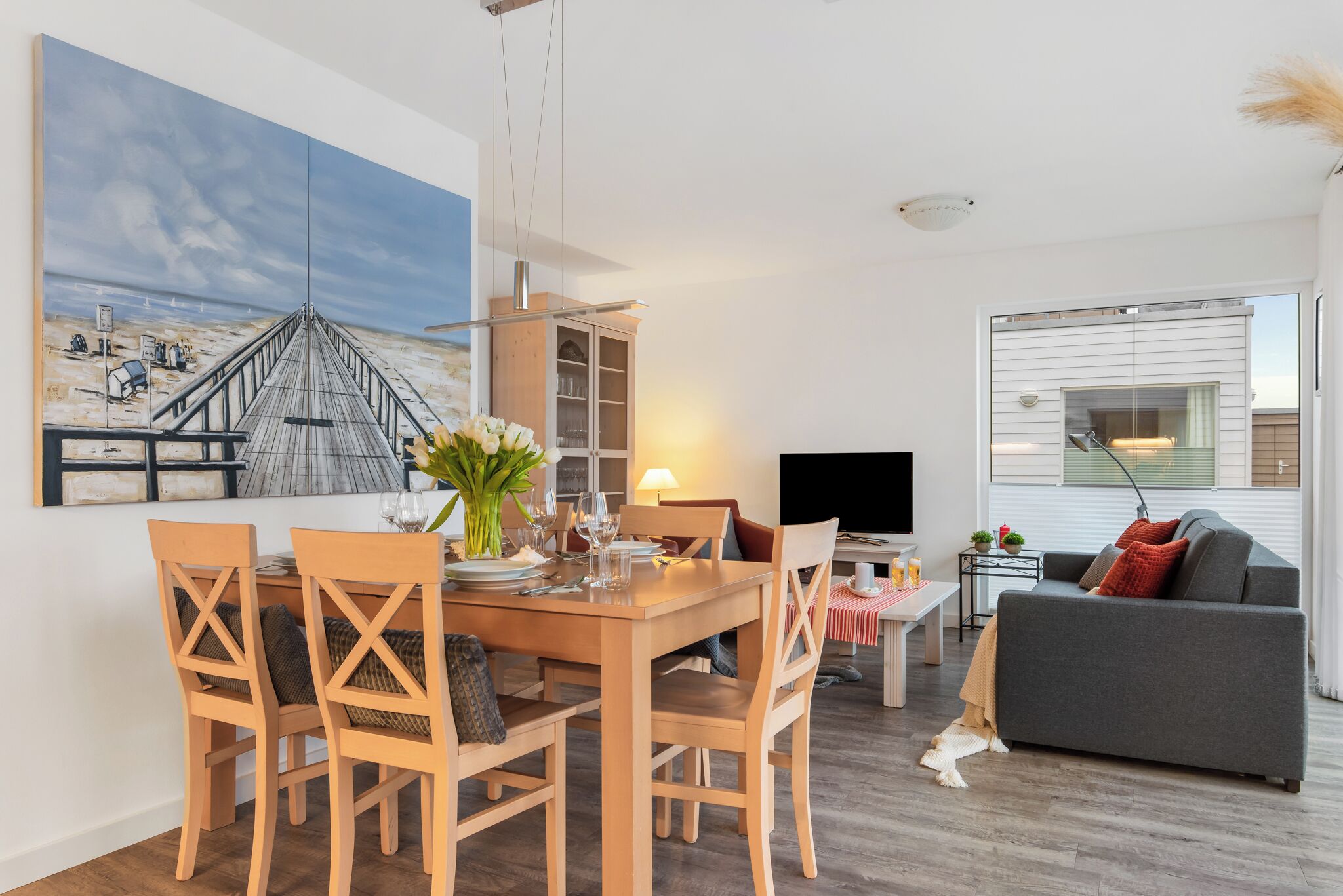 Ett modernt vardagsrum med ett matbord i trä för fyra personer, en grå soffa och en stor tavla som föreställer en brygga.