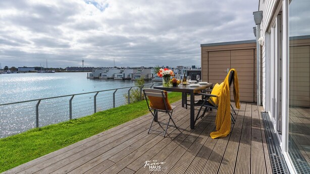 Eine Terrasse mit Tisch für zwei Personen, mit Blick auf ein Gewässer, im Hintergrund Boote und Gebäude.