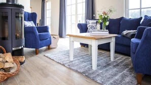 A living room with a black wood-burning stove, blue upholstered chairs, a white coffee table, and a large window with curtains. - Luxushaus Boddenkuste mit Sauna (Fuhlendorf)